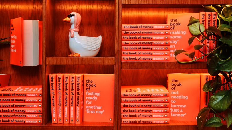 Orange books on a bookshelf. The book's title is "The book of Money" the author is Monzo. There is also a duck statue. The duck has sunglasses and a scarf over its head. 