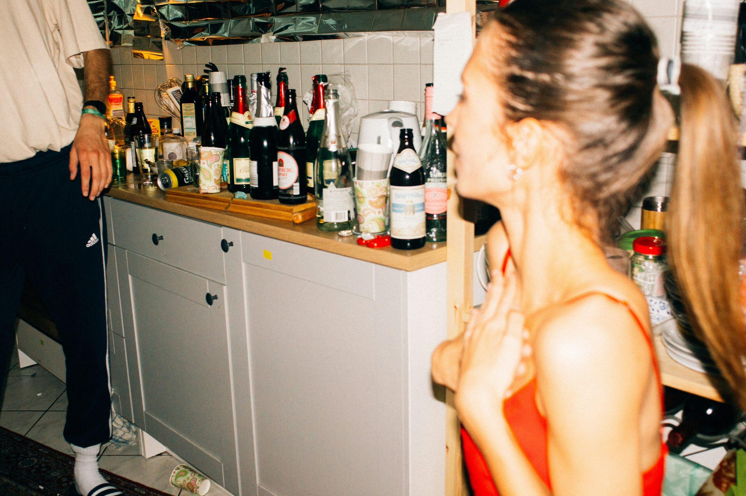 A party scene, showing a kitchen counter crammed with open bottles and cans - with out of focus people.